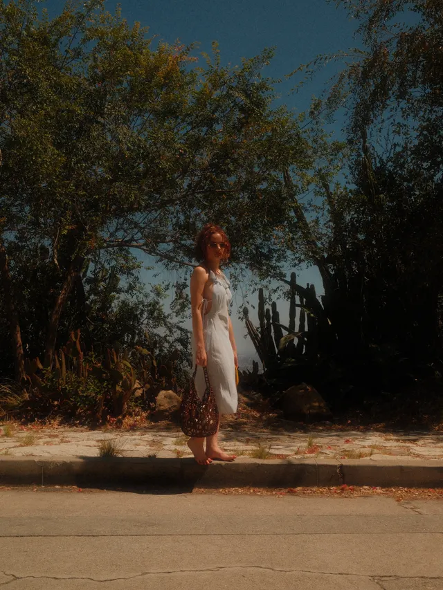 Joey King standing barefoot on a kerb in a pale dress.