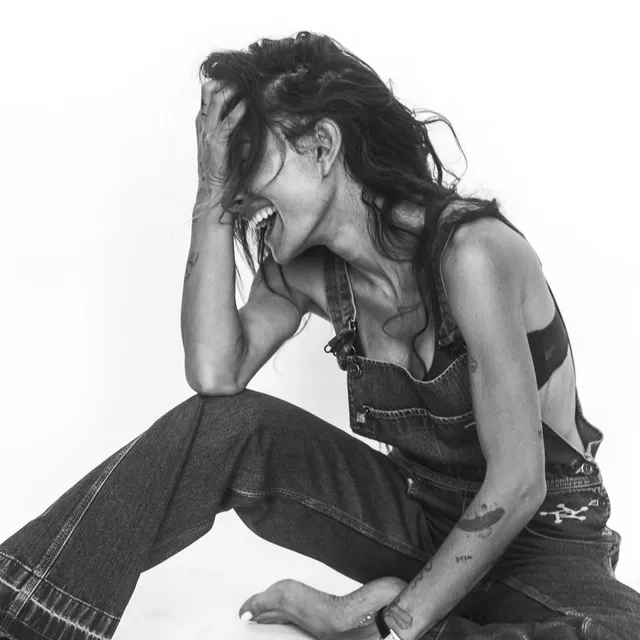 Sarah Shahi sitting for a black-and-white portrait in denim overalls.