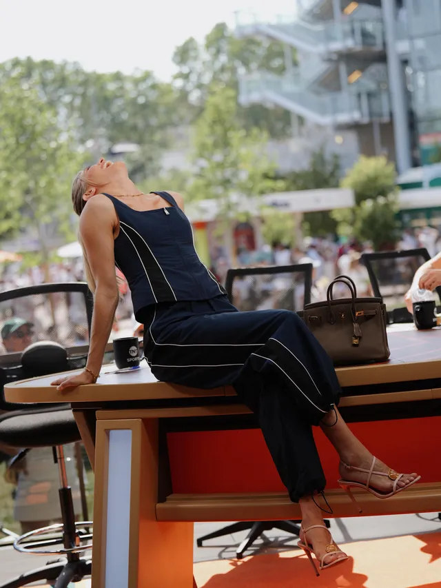 Eugenie Bouchard sitting on a table in heeled sandals and a dark sleeveless outfit.