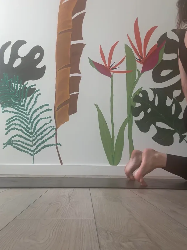 Alexandra Daddario extending a bare foot beside a tropical wall mural.