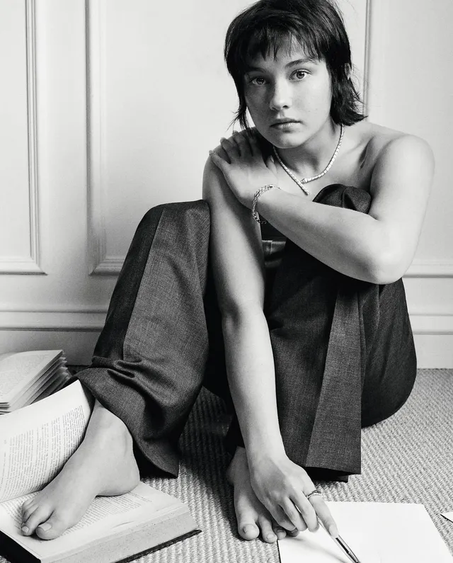 Cailee Spaeny sitting barefoot beside an open book in a monochrome portrait.