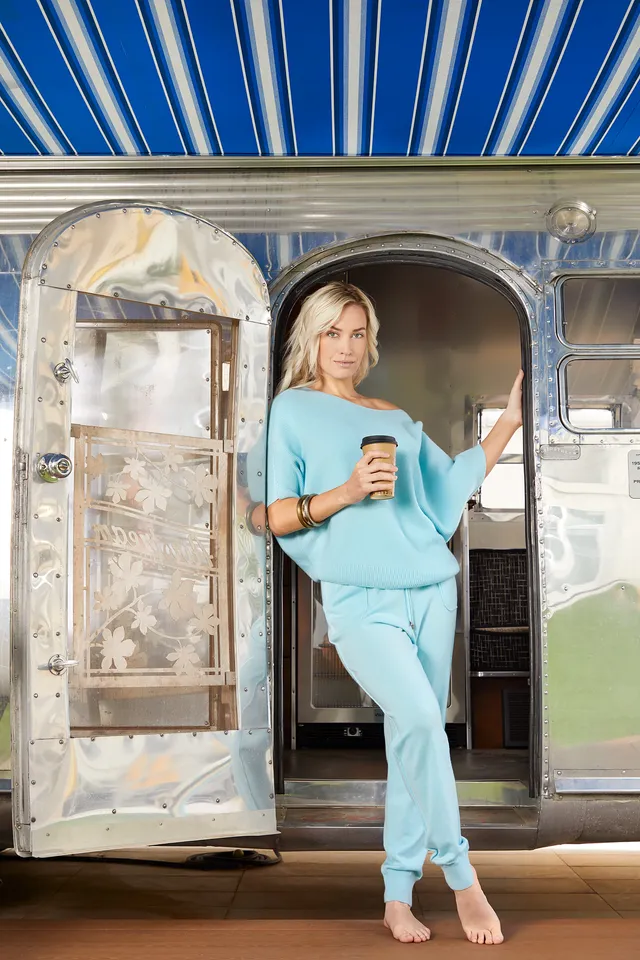 Paige Spiranac standing barefoot in a turquoise outfit in a trailer doorway.