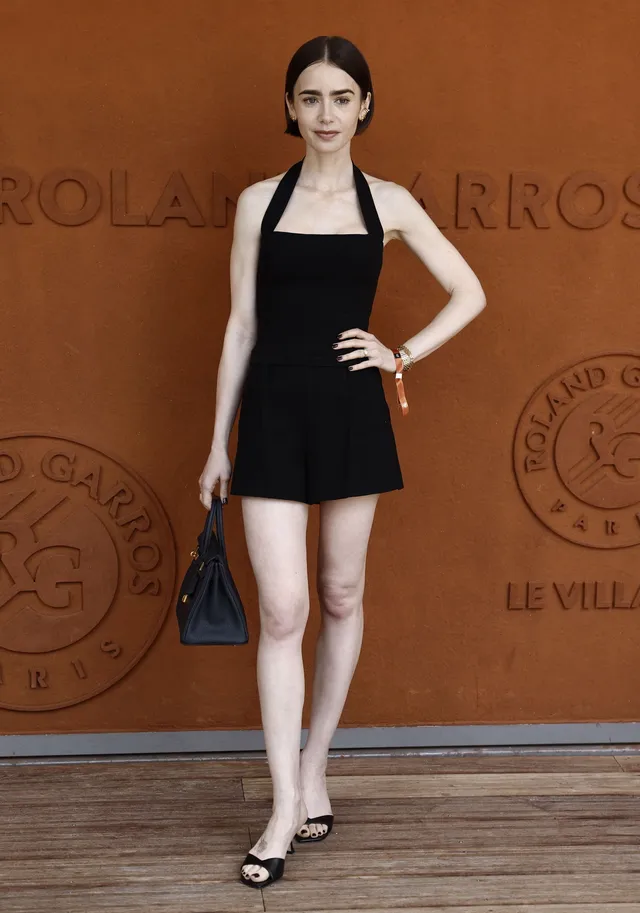 Lily Collins standing at an event in a black dress and heels.