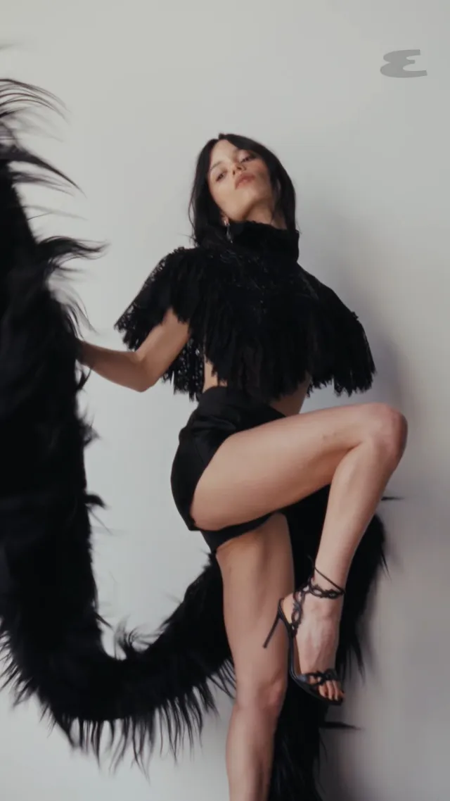 Jenna Ortega: seated in a black feathered outfit and heels.
