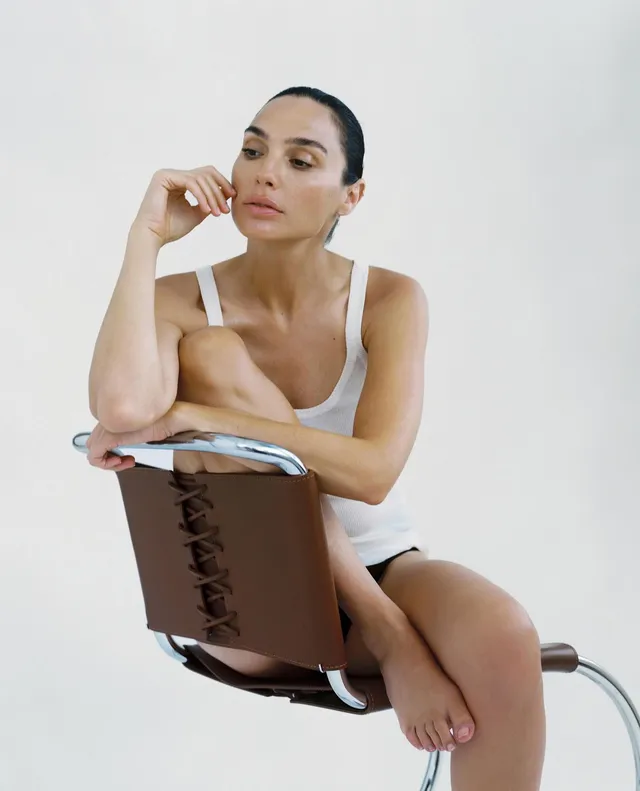 Gal Gadot sitting barefoot on a chair in a studio portrait.