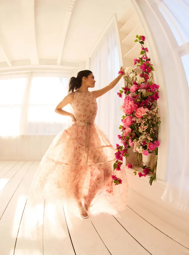 Rachel Zegler posing beside flowers in a flowing pale pink dress.