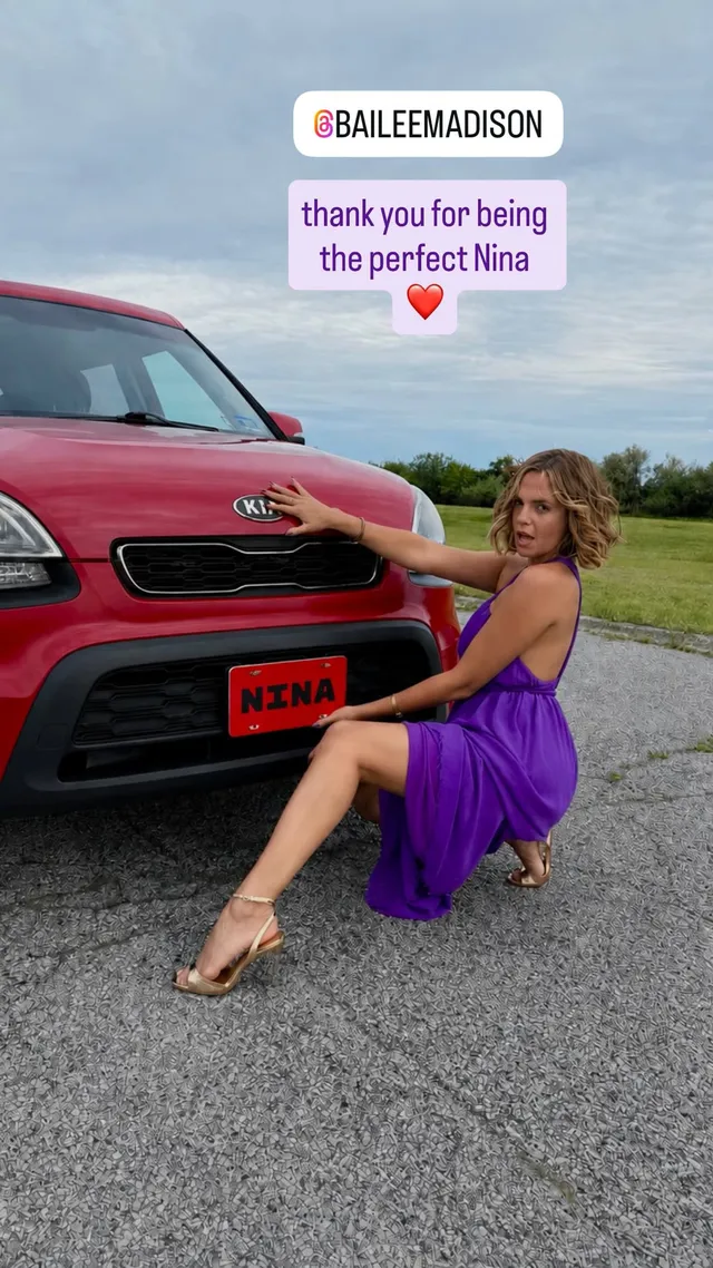 Bailee Madison crouching beside a red vehicle in a purple dress and clear heels.