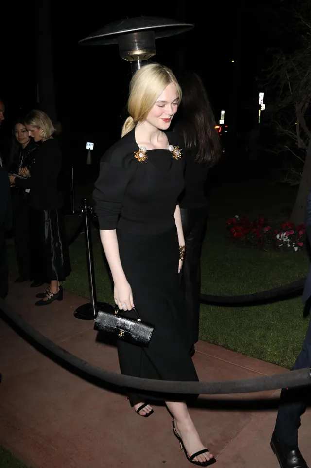 Elle Fanning walking outside at night in a black dress and heeled sandals.