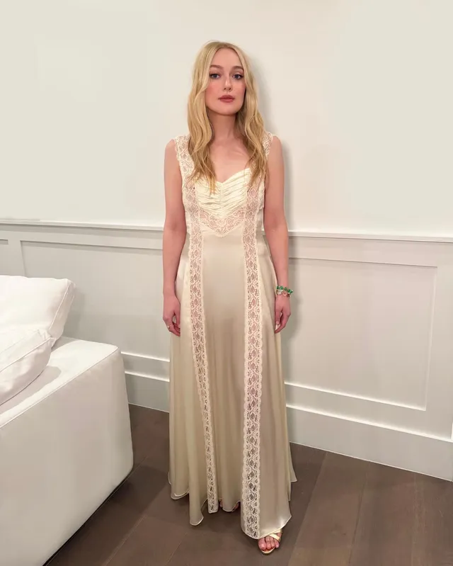 Dakota Fanning posing indoors in a long cream dress and sandals.