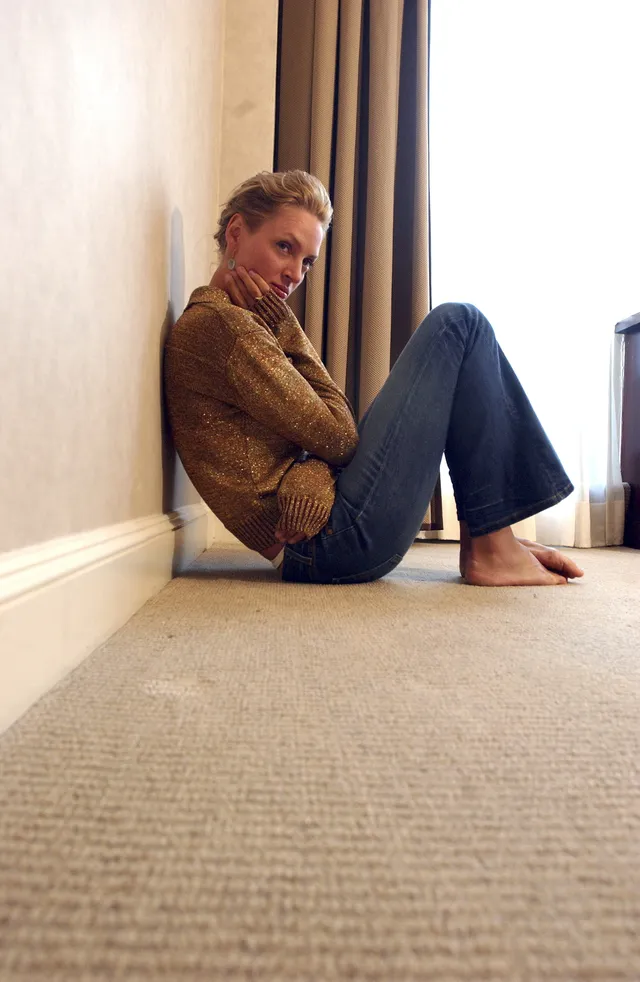 Uma Thurman sitting barefoot against a wall in jeans.