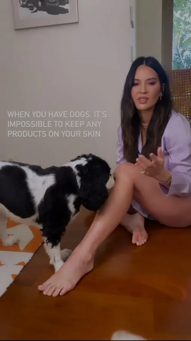 Olivia Munn sitting barefoot beside a dog on a wooden floor.