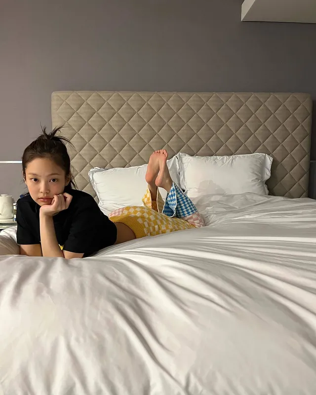 Jennie Kim lying on a bed in a dark top with her bare feet raised.