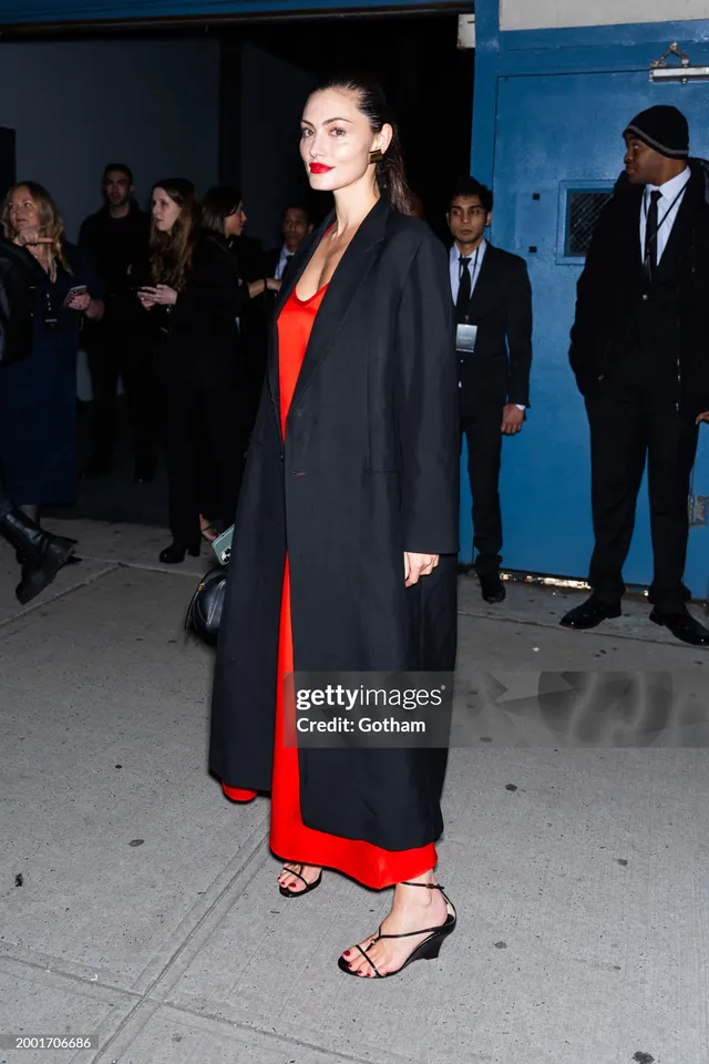 Phoebe Tonkin walking outside in a red dress and black coat.