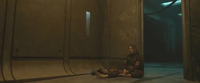 Rebecca Ferguson sitting barefoot on a floor beside kitchen cabinets in a film scene.