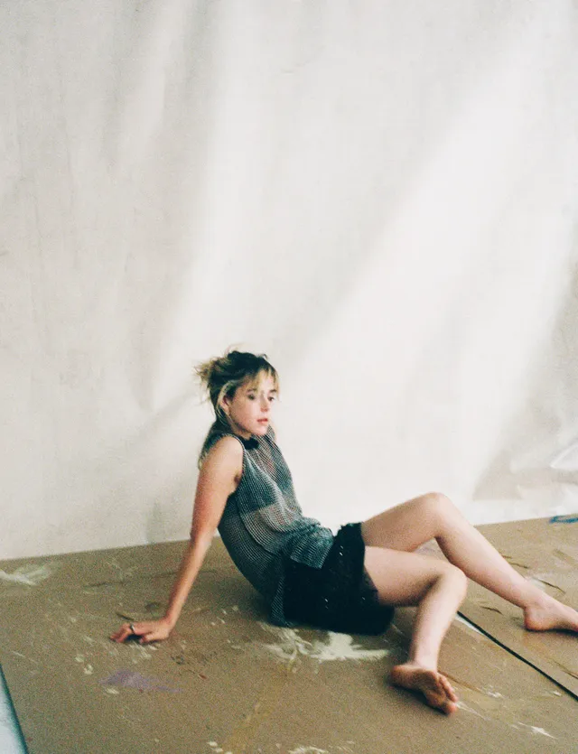 Kiernan Shipka sitting barefoot on a wooden platform in a dark dress.