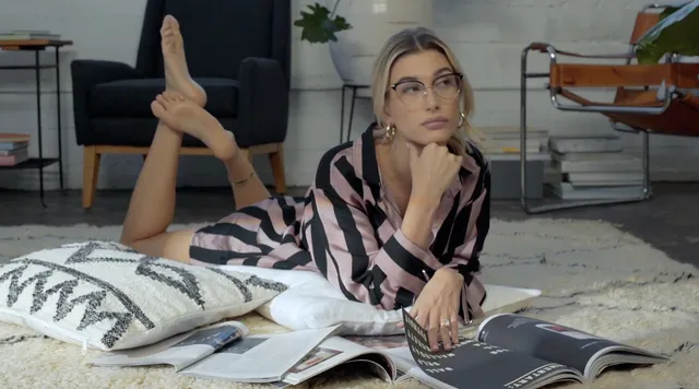 Hailey Bieber reading on a rug with bare feet raised.