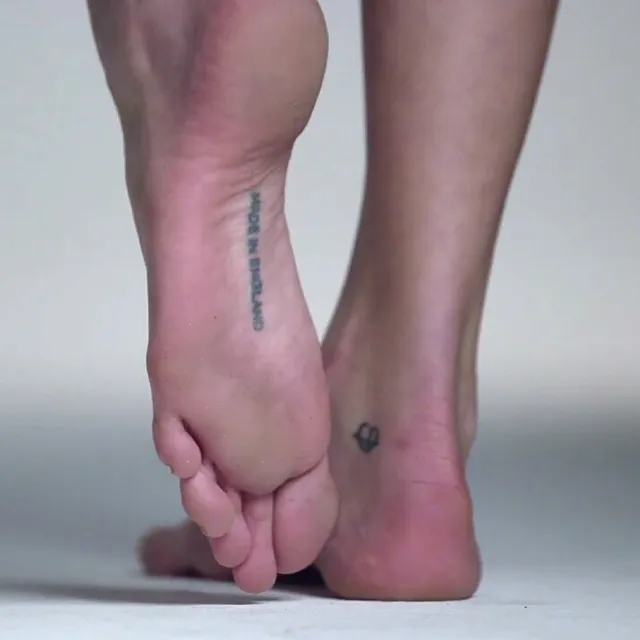 Cara Delevingne standing on tiptoe with a foot tattoo visible.