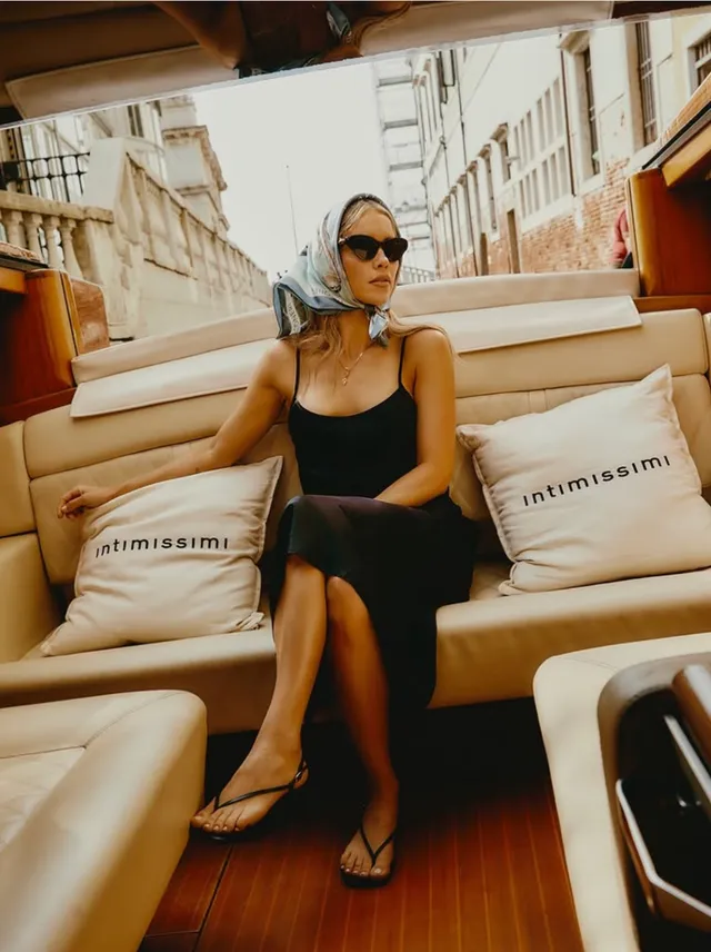 Claire Holt sitting on a boat in a black dress and flip-flops.