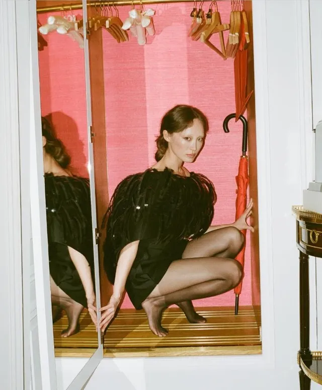 Havana Rose Liu crouching in a black dress and gold heels beside a mirror.