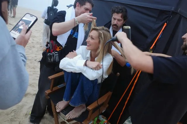 Bella Hadid seated barefoot in a chair while stylists work nearby.
