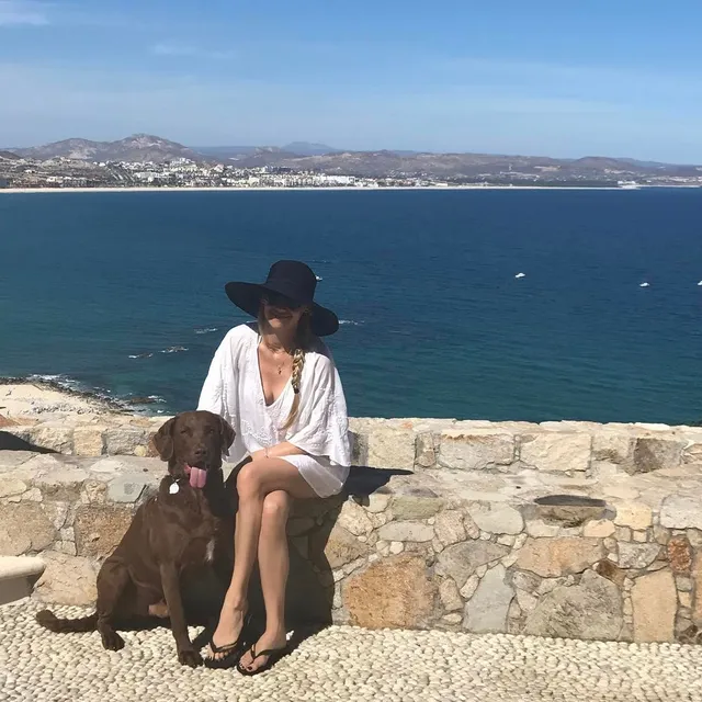 Anna Kournikova sitting beside a dog in flat sandals overlooking the coast.
