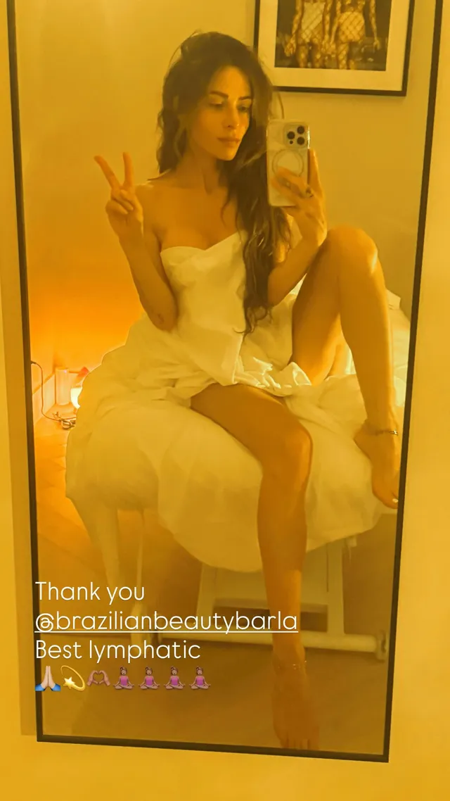 Sarah Shahi taking a seated mirror portrait in a pale outfit.