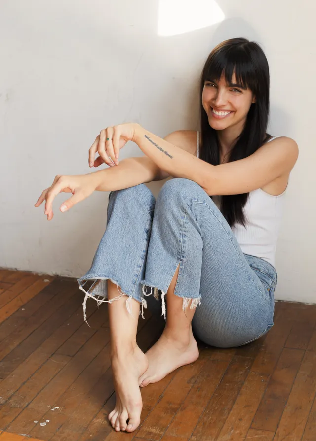 María Gabriela de Faría sitting barefoot on a wooden floor in jeans.