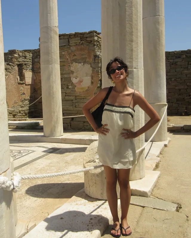 Camila Cabello standing by stone columns in a pale dress and sandals.