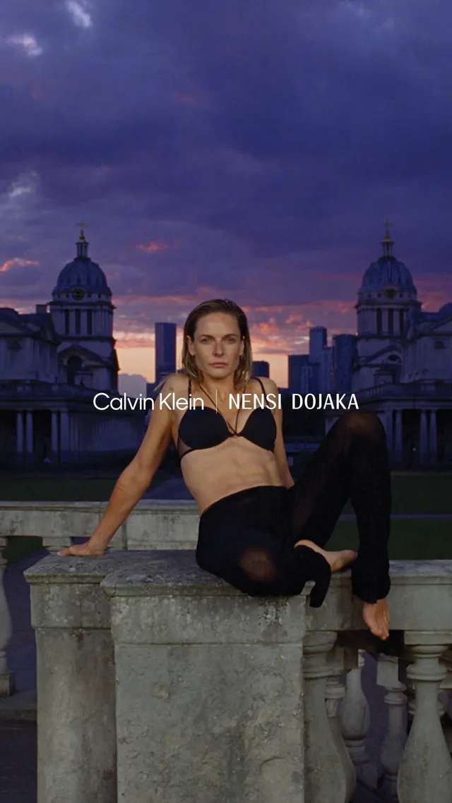 Rebecca Ferguson posing barefoot in a dark outfit beside a stone railing at dusk.