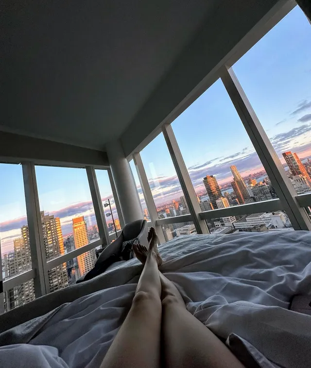 Úrsula Corberó reclining on a bed with bare legs crossed in front of a window.
