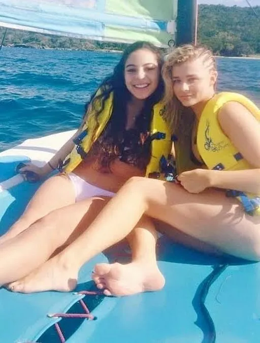 Chloë Grace Moretz sitting barefoot on a boat in a yellow life jacket beside another person.