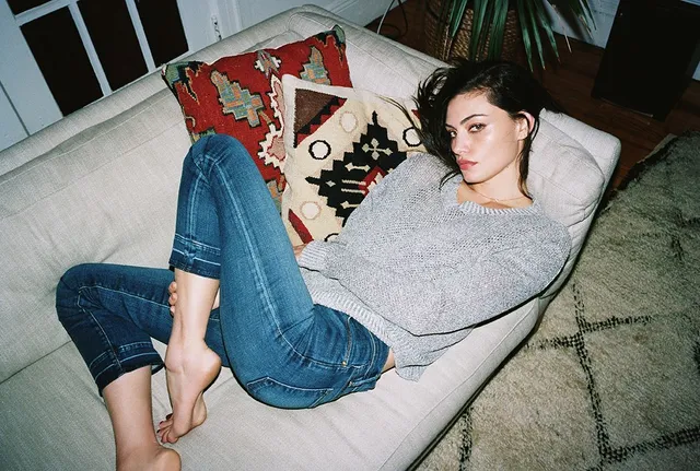 Phoebe Tonkin reclining on a sofa in jeans and a sweater.