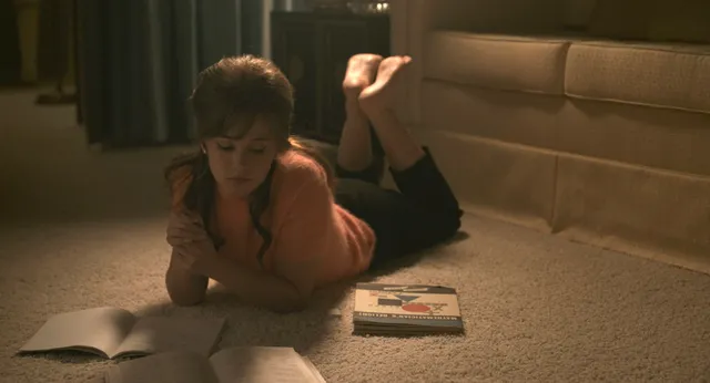 Cailee Spaeny reading while lying barefoot on the floor in a film scene.