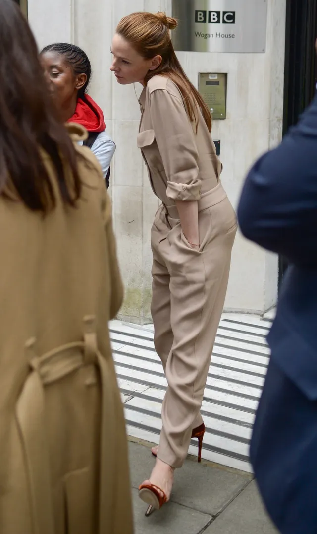Rebecca Ferguson standing in a beige jumpsuit and open-toe heels during an interview.