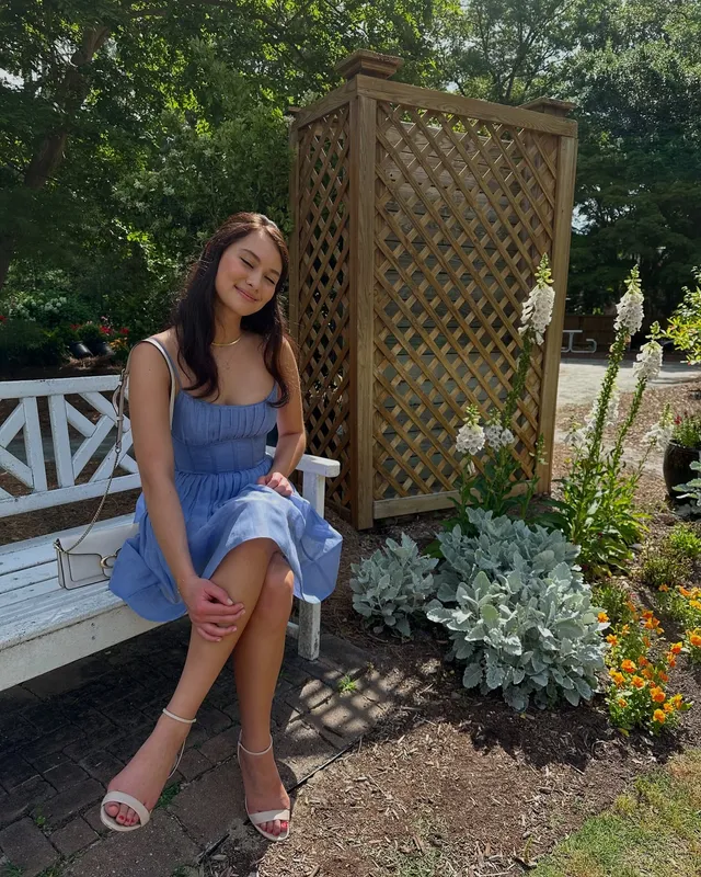 Lola Tung sitting on a garden bench in a blue dress and white heels.