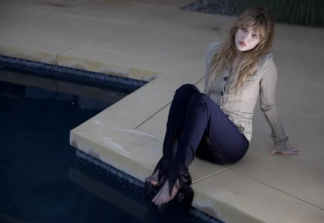 Sophie Thatcher sitting beside a pool in dark trousers and heeled sandals.