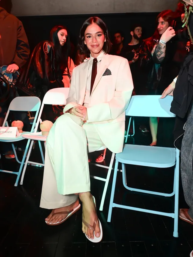 Bruna Marquezine sitting at an event in a pale suit and flat sandals.