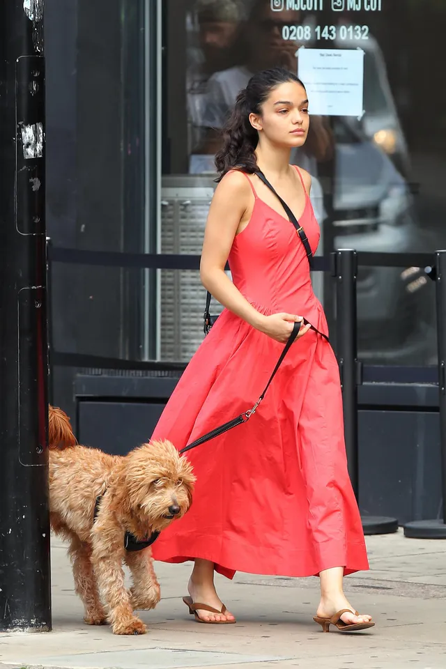 Rachel Zegler walking a dog in a red dress and flat sandals.