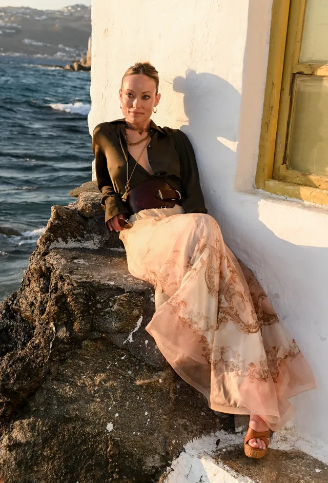 Olivia Wilde sitting beside the sea in a long skirt and platform sandals.