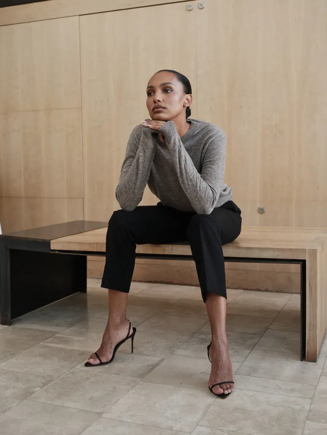 Jasmine Tookes sitting on a bench in black heels, cropped trousers and a grey sweater.