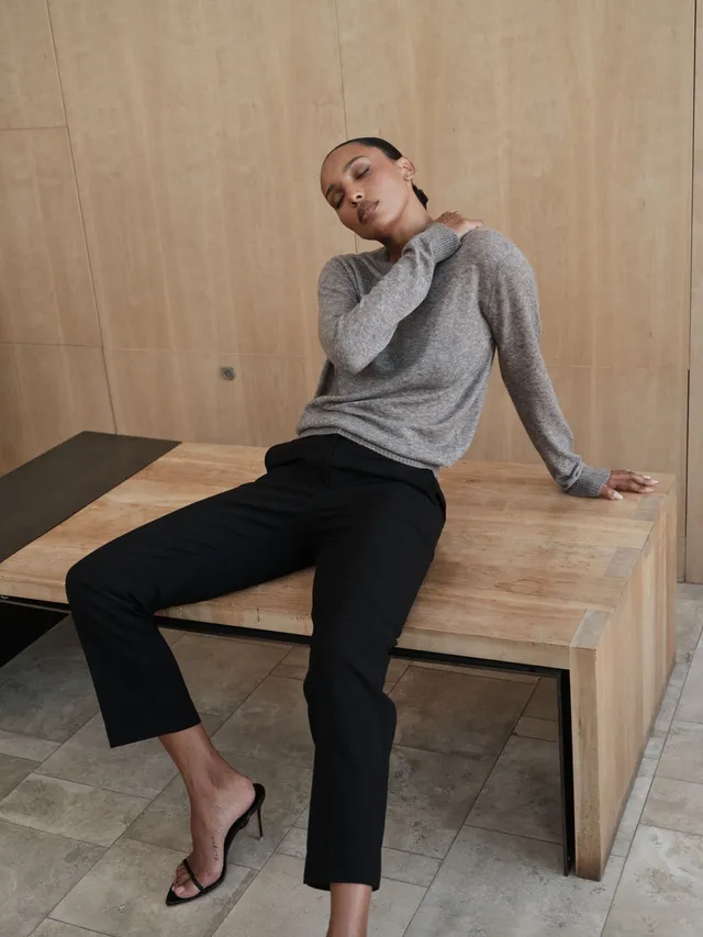 Jasmine Tookes sitting on a wooden table in black heels and a grey sweater.