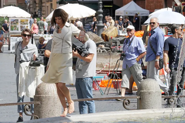 Úrsula Corberó balancing barefoot along a stone fountain edge in a pale dress.