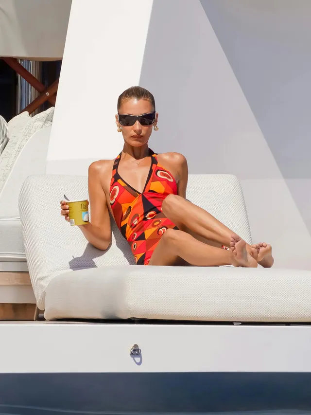Bella Hadid sitting barefoot on a boat lounger in a colorful dress.