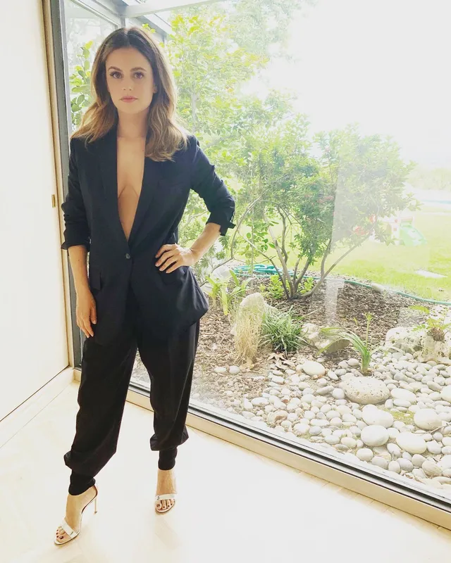 Rachel Bilson standing by a window in a dark tailored outfit.