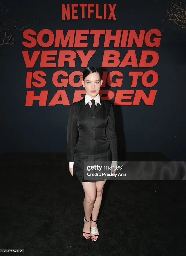 Victoria Pedretti standing beside a film poster in a dark outfit.