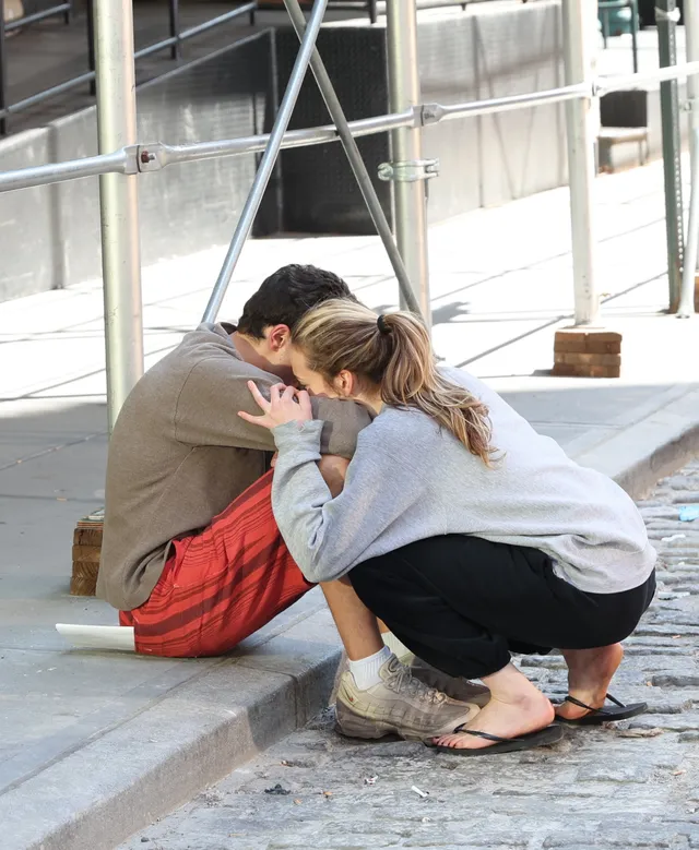 Sarah Pidgeon crouching beside a man outdoors in a grey top and flat sandals.