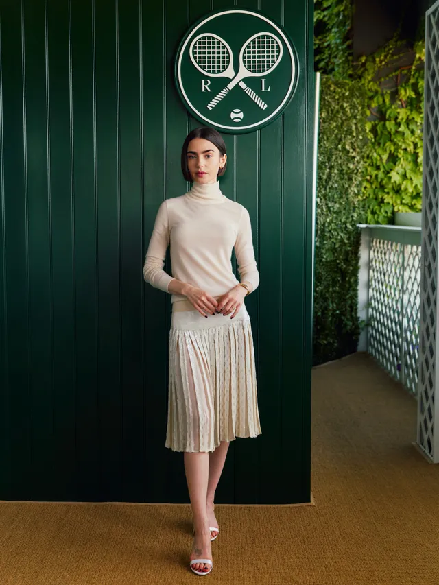 Lily Collins wearing a pleated skirt and pale heels indoors.