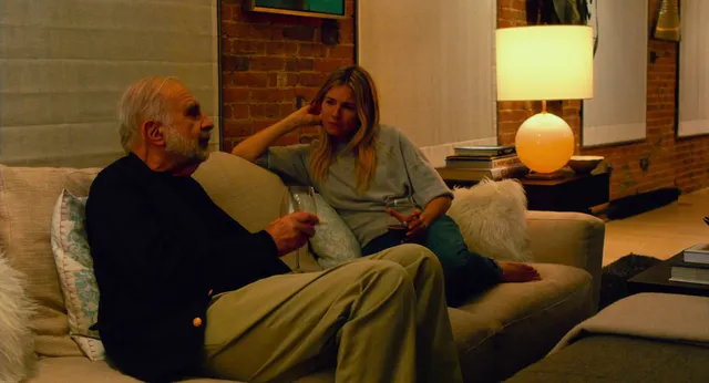 Sienna Miller sitting beside a man on a sofa in a screen scene.