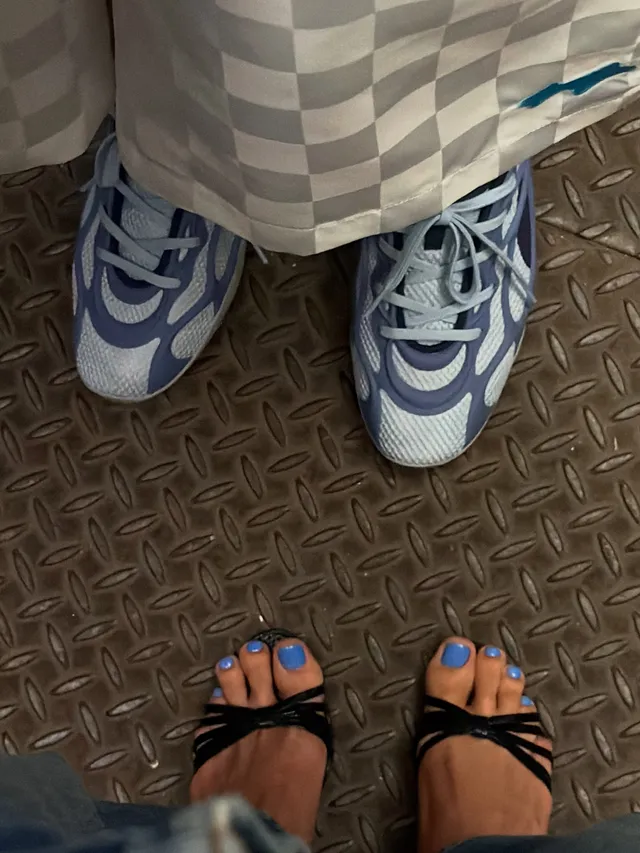 Hailey Bieber wearing black strappy heels beside another person’s trainers.