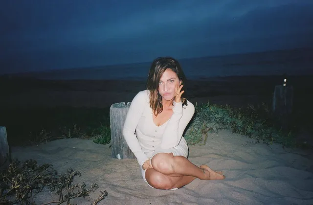 Brittany Snow sitting on sand in a pale sweater.